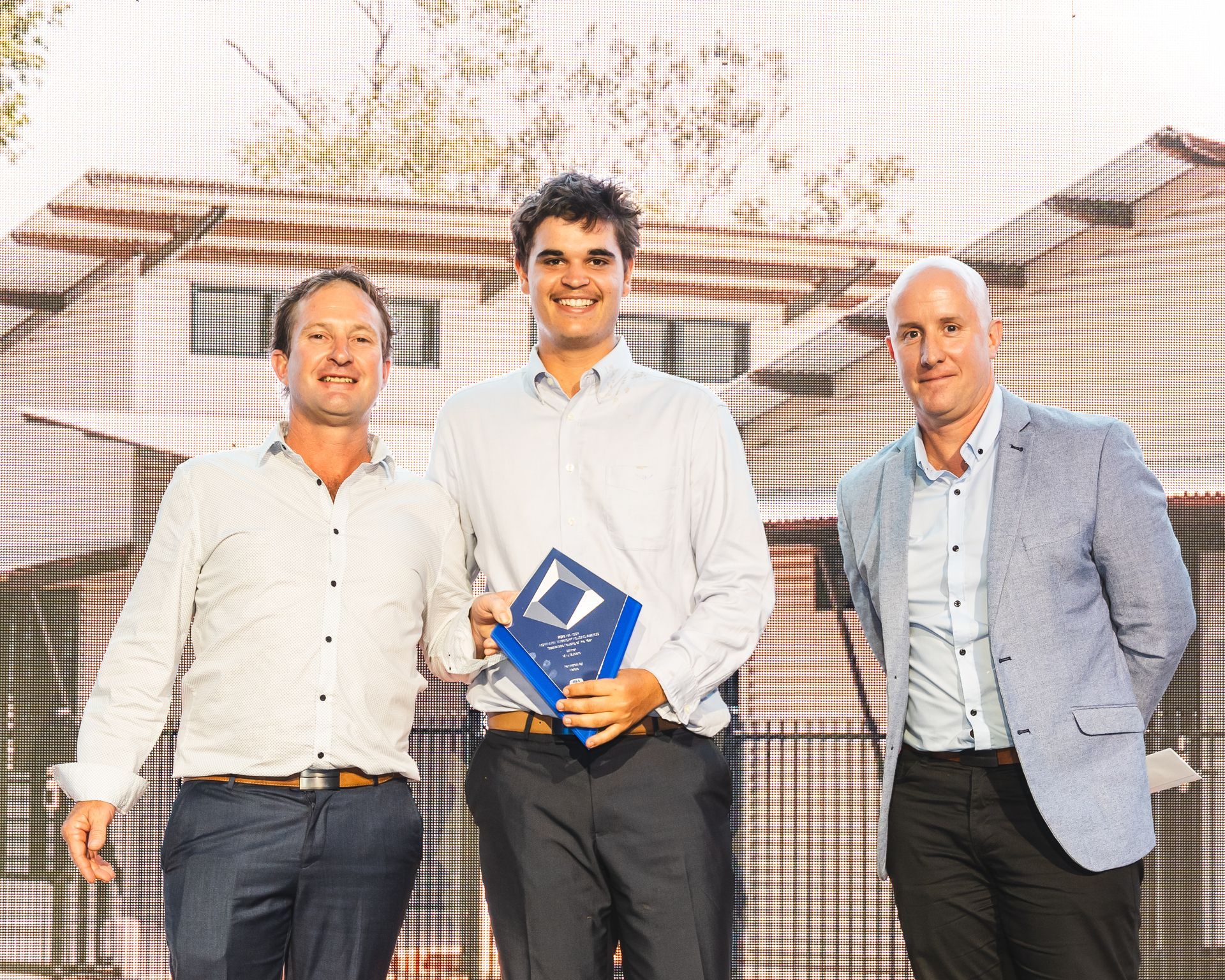 Three men at an awards event; the center man holding a trophy, smiling.