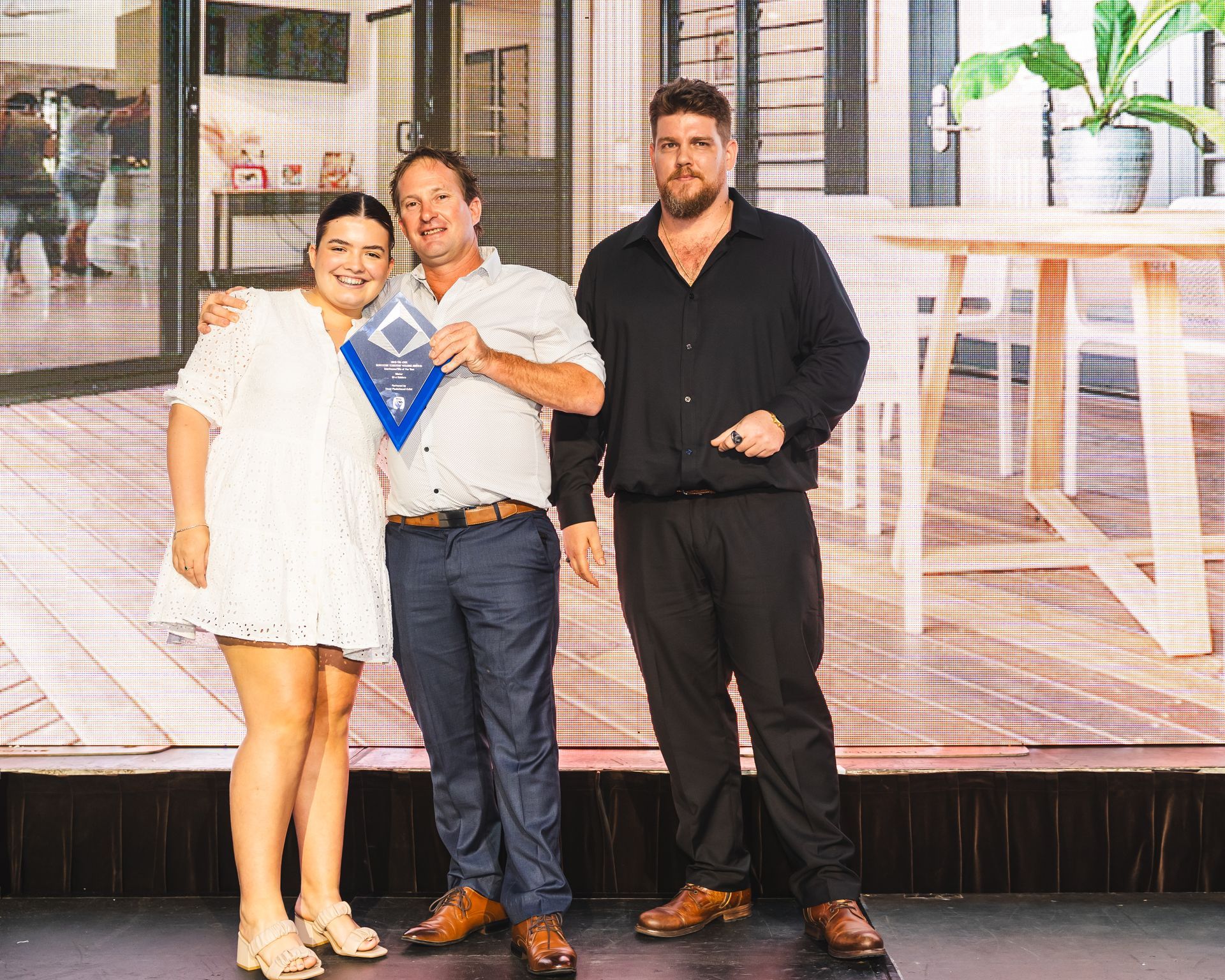 Three people smiling, holding an award plaque, inside modern building.