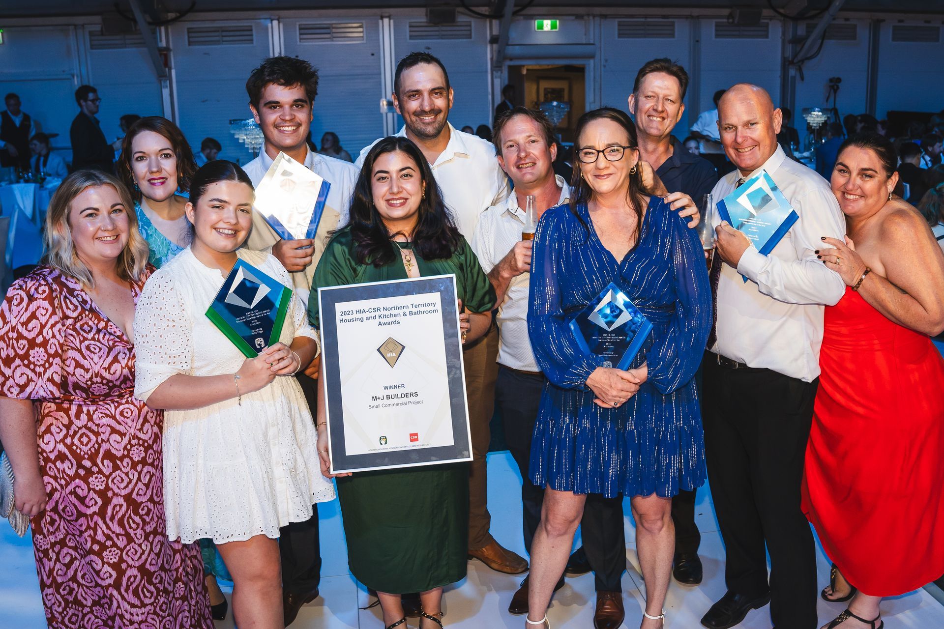 Group of people at an awards ceremony, holding trophies and a certificate. Cheerful expressions, some in formal wear, in a hall.