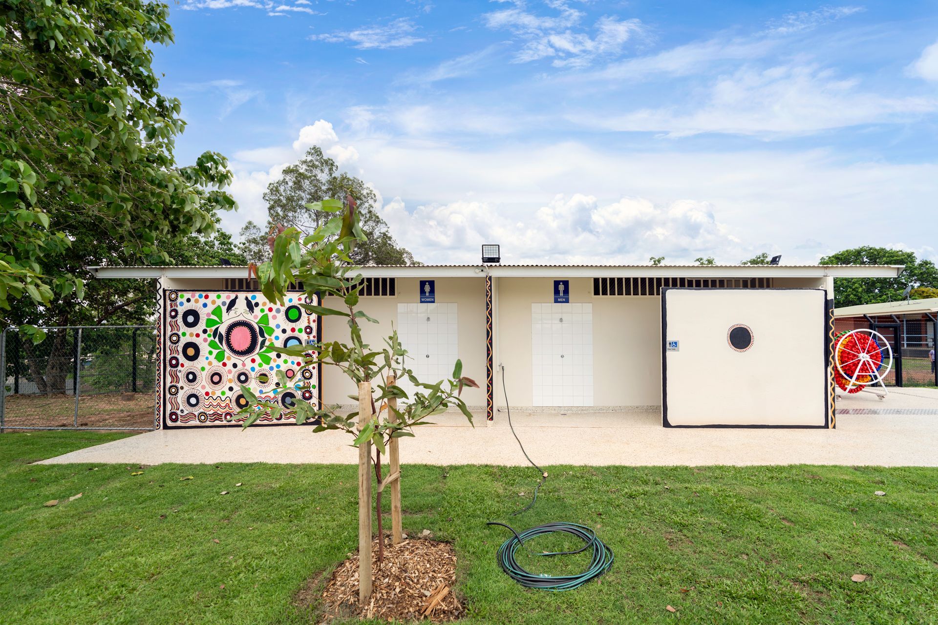 Public restrooms with colorful mural art, set on green grass with a tree under a blue sky.