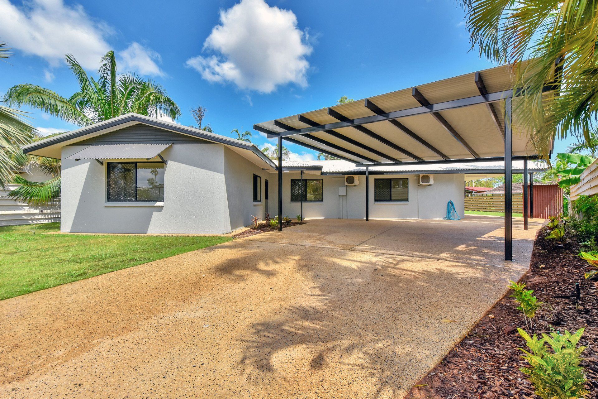 A light-colored house with a carport. Driveway and lawn. Sunny, cloudy day.