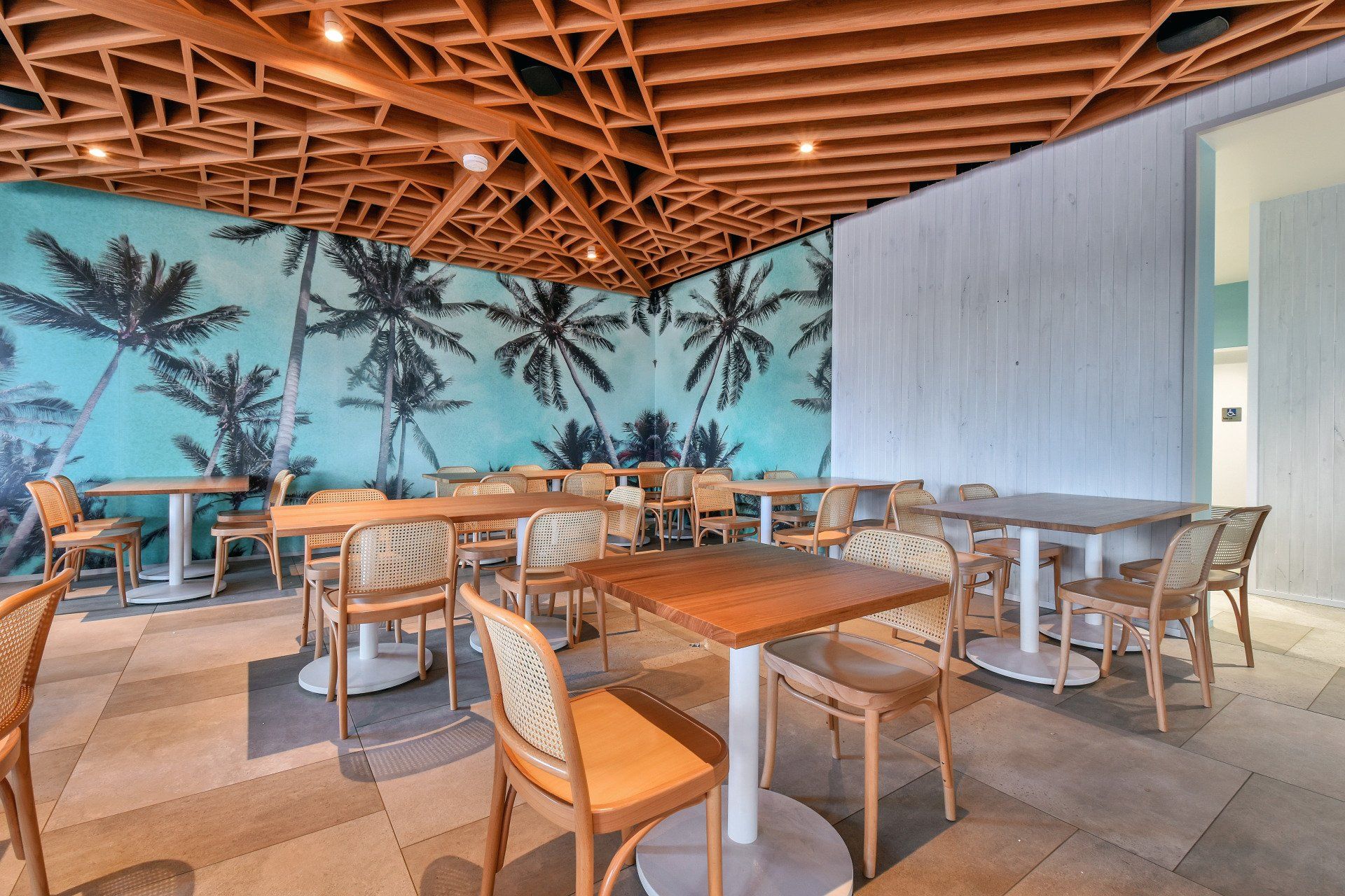 Cafe interior with palm tree mural, wooden tables, woven chairs, and intricate ceiling design.