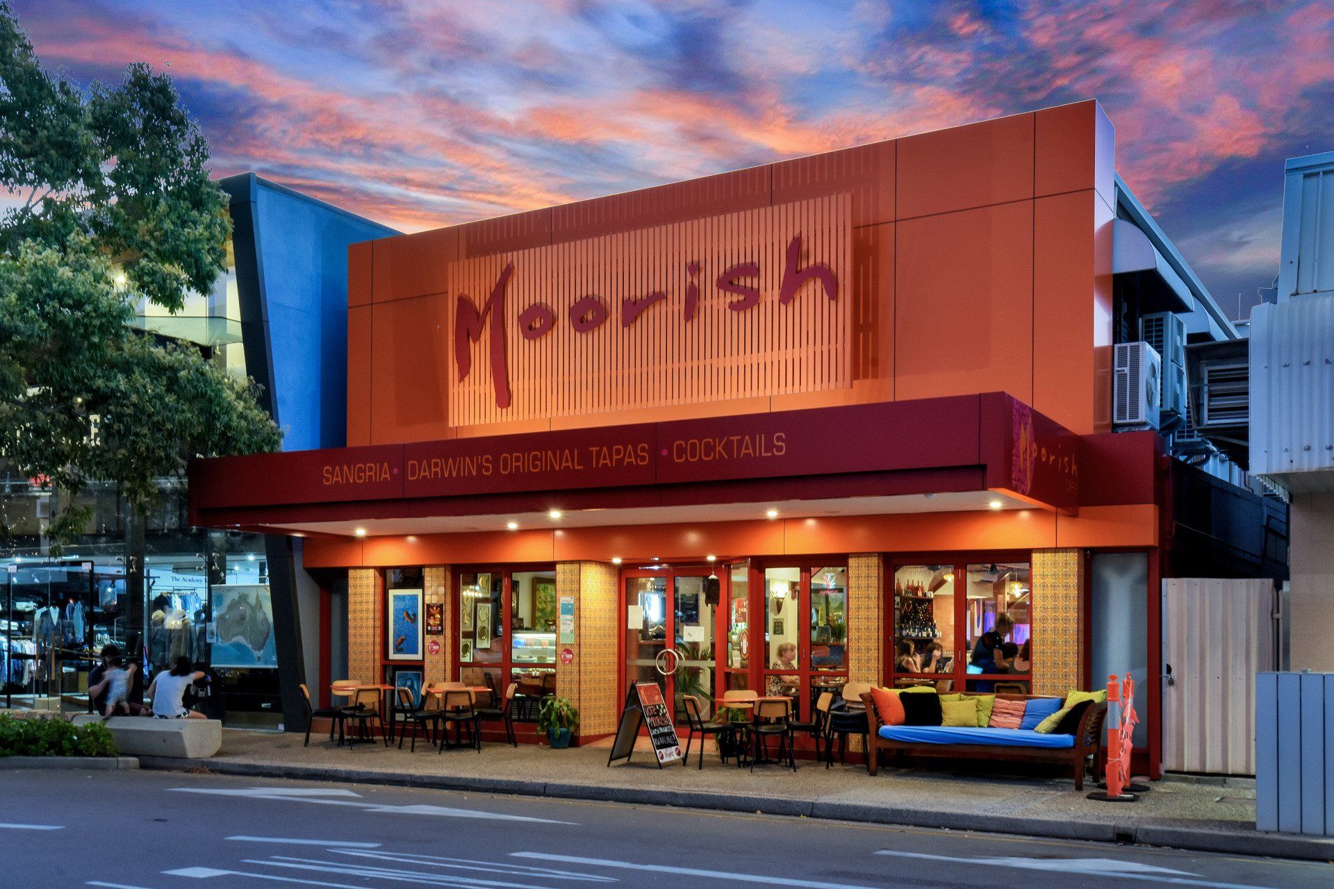 Moorish Tavern exterior with orange facade, tables, and people. Evening sky.