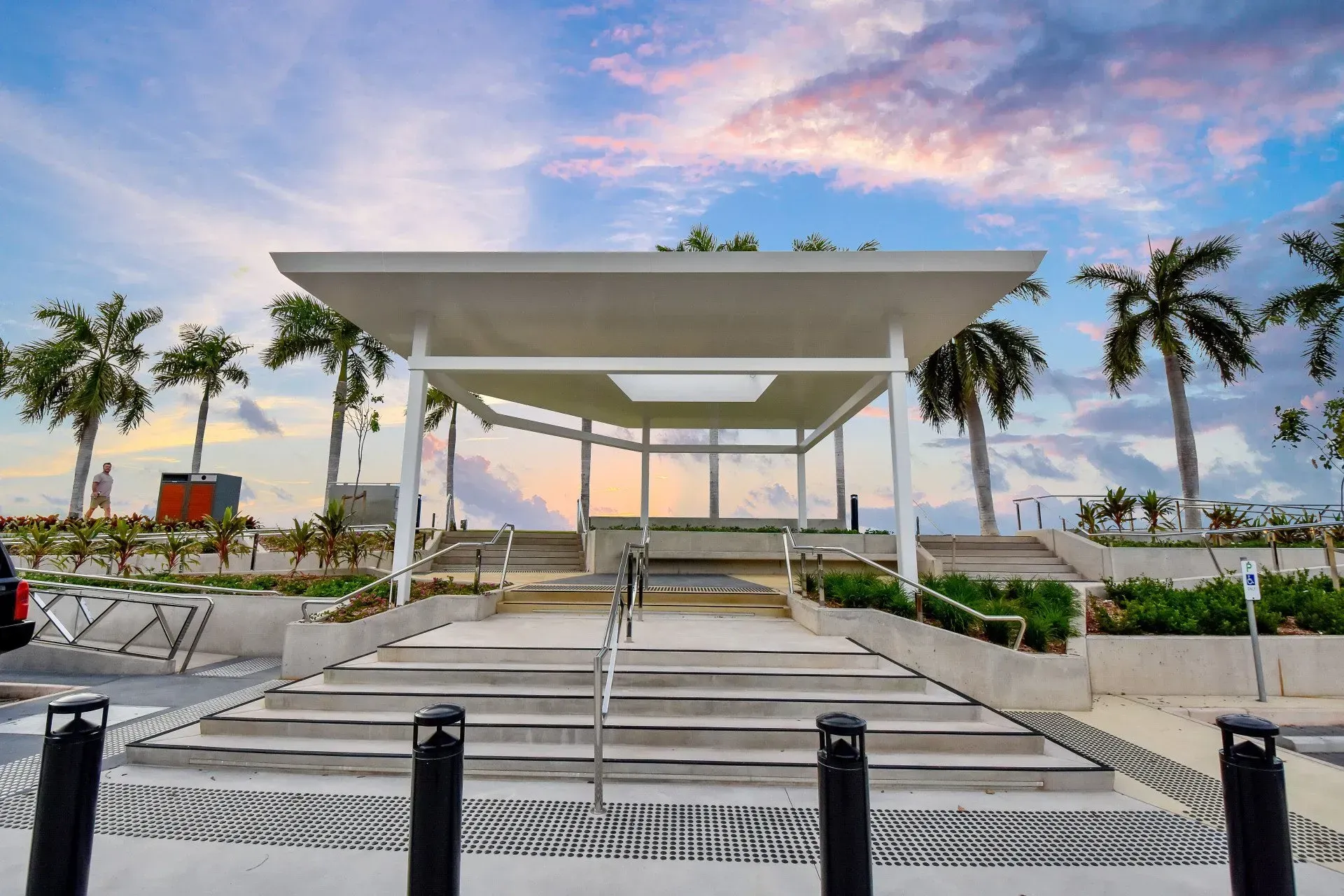 A modern pavilion with steps leading up, palm trees, and a sunset sky.