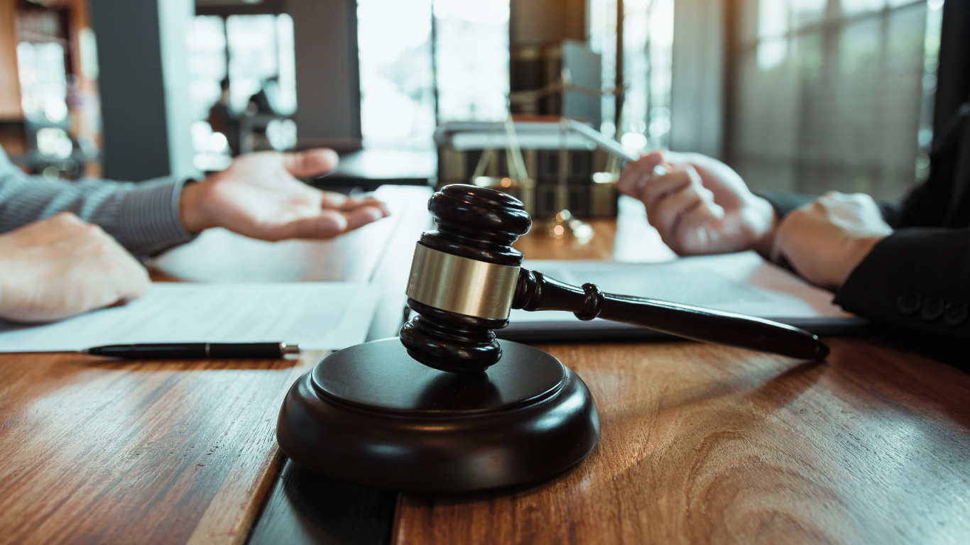 Gavel on wooden table with two people and documents, likely in a courtroom.