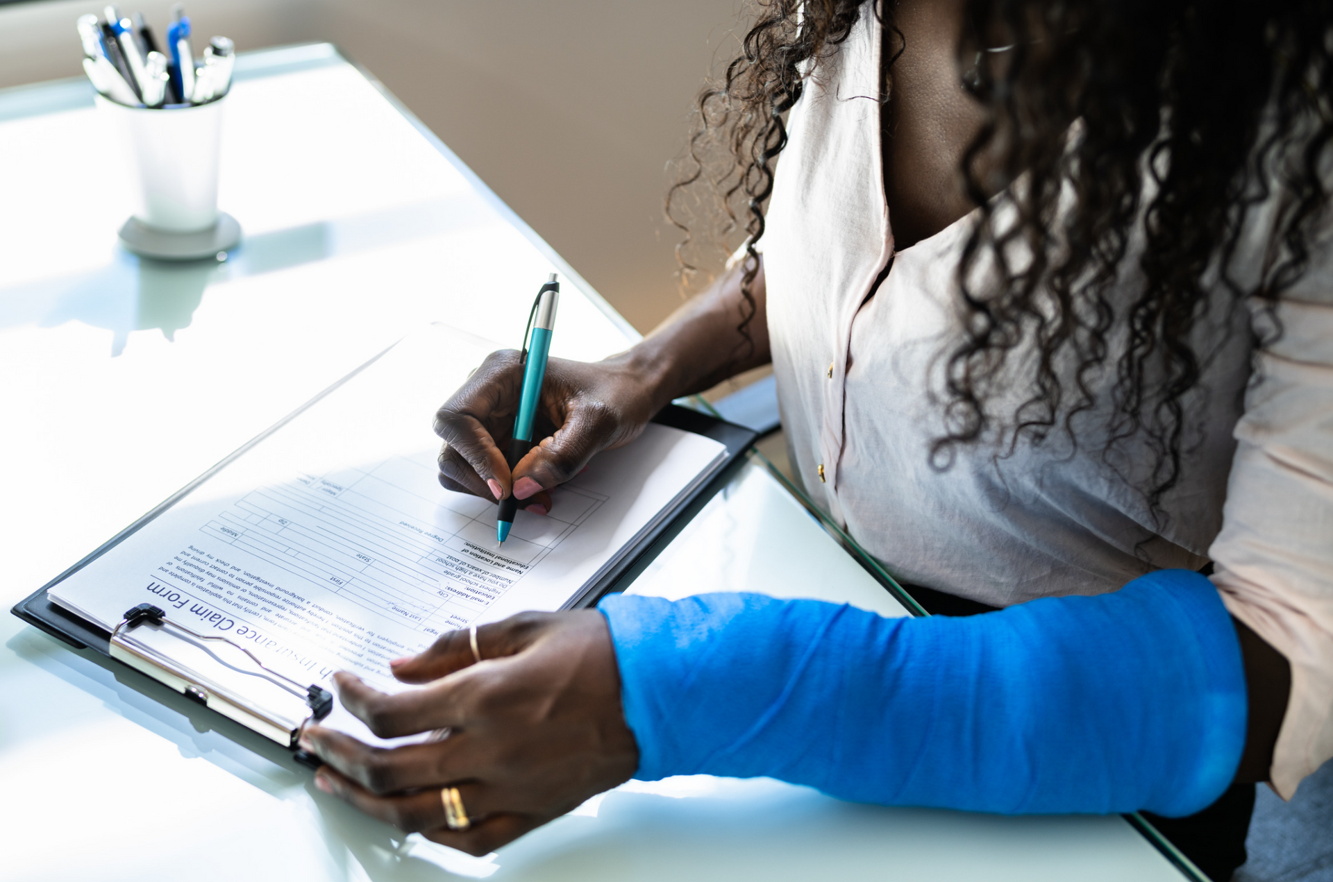Person with a blue cast writing on a clipboard at a desk.