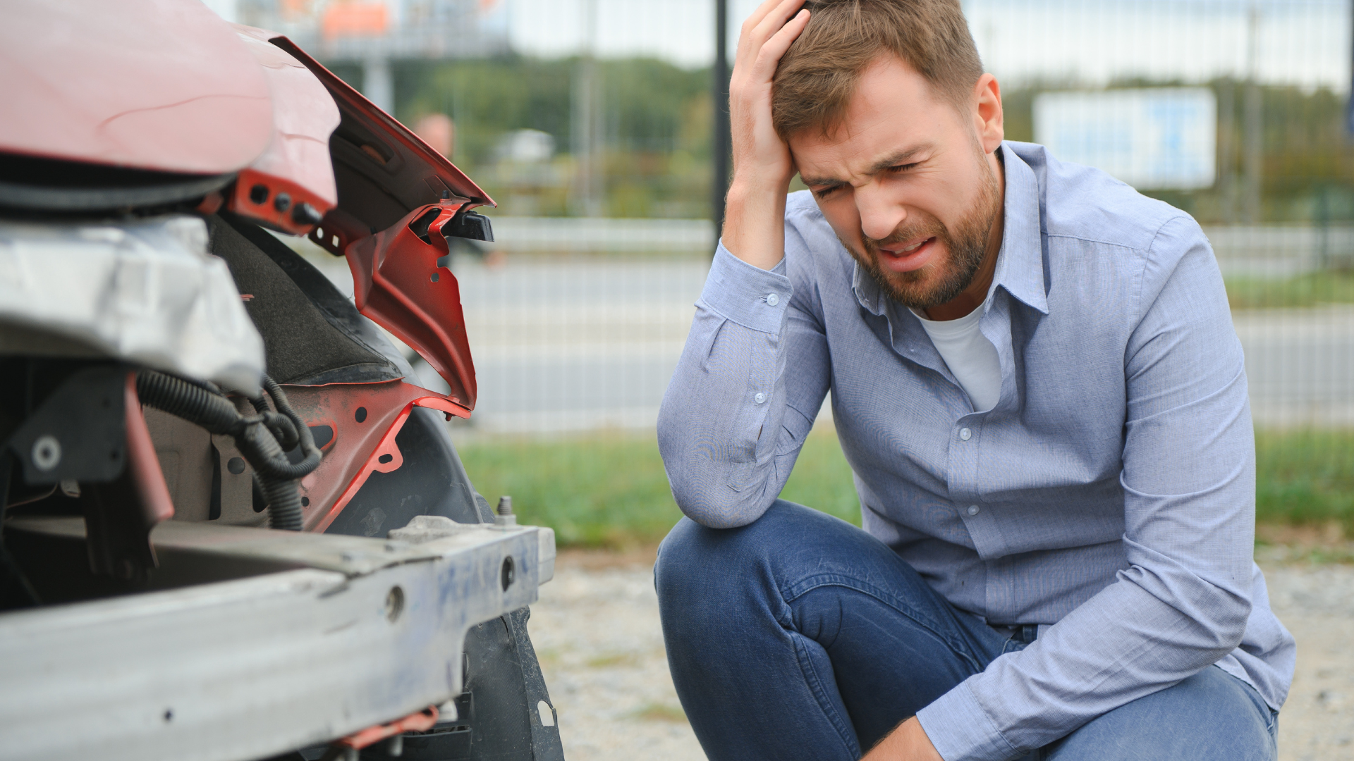 Man kneels near damaged red car, holding his head, looking distressed. Outdoor setting.