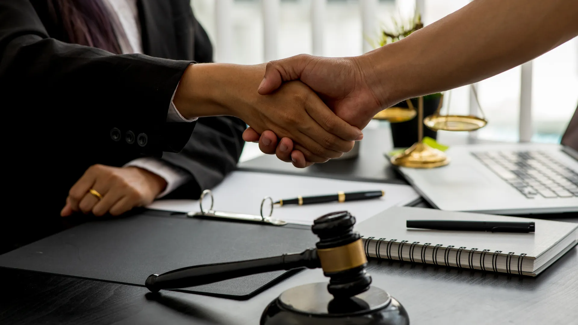 Two people shaking hands over a desk with a gavel, legal documents, and a scale of justice.