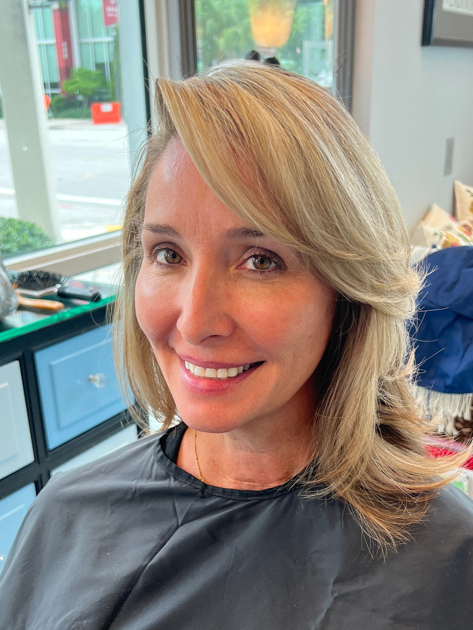 Woman with blonde layered hair smiles, possibly at a hair salon.