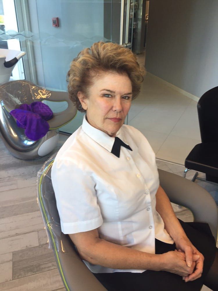Woman with short, curly hair, in a white shirt with a black bow tie, sitting in a modern chair.