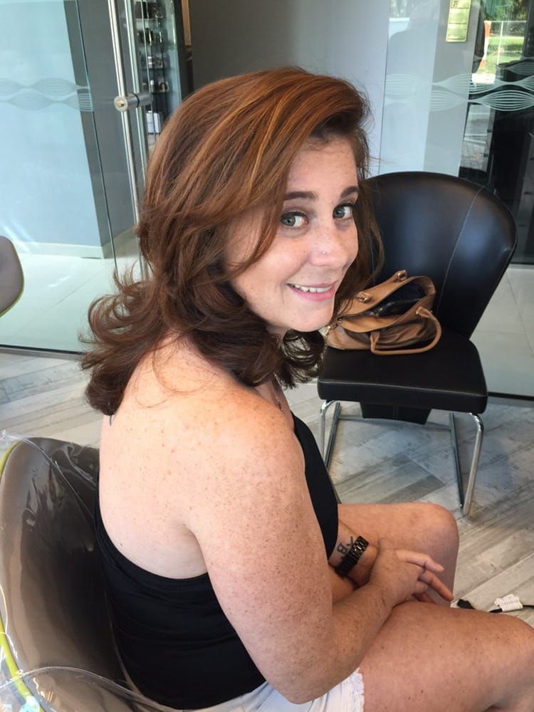 Woman with auburn hair smiles in a salon chair. She wears a black top and white shorts.