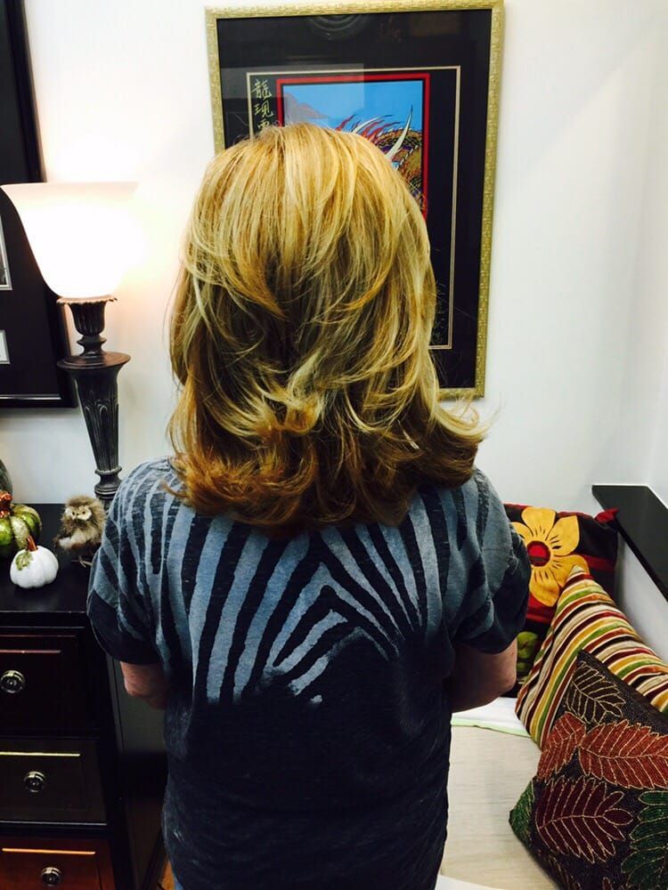 Woman with layered blonde hair, wearing gray sweater, standing in a room with a lamp and artwork on the wall.