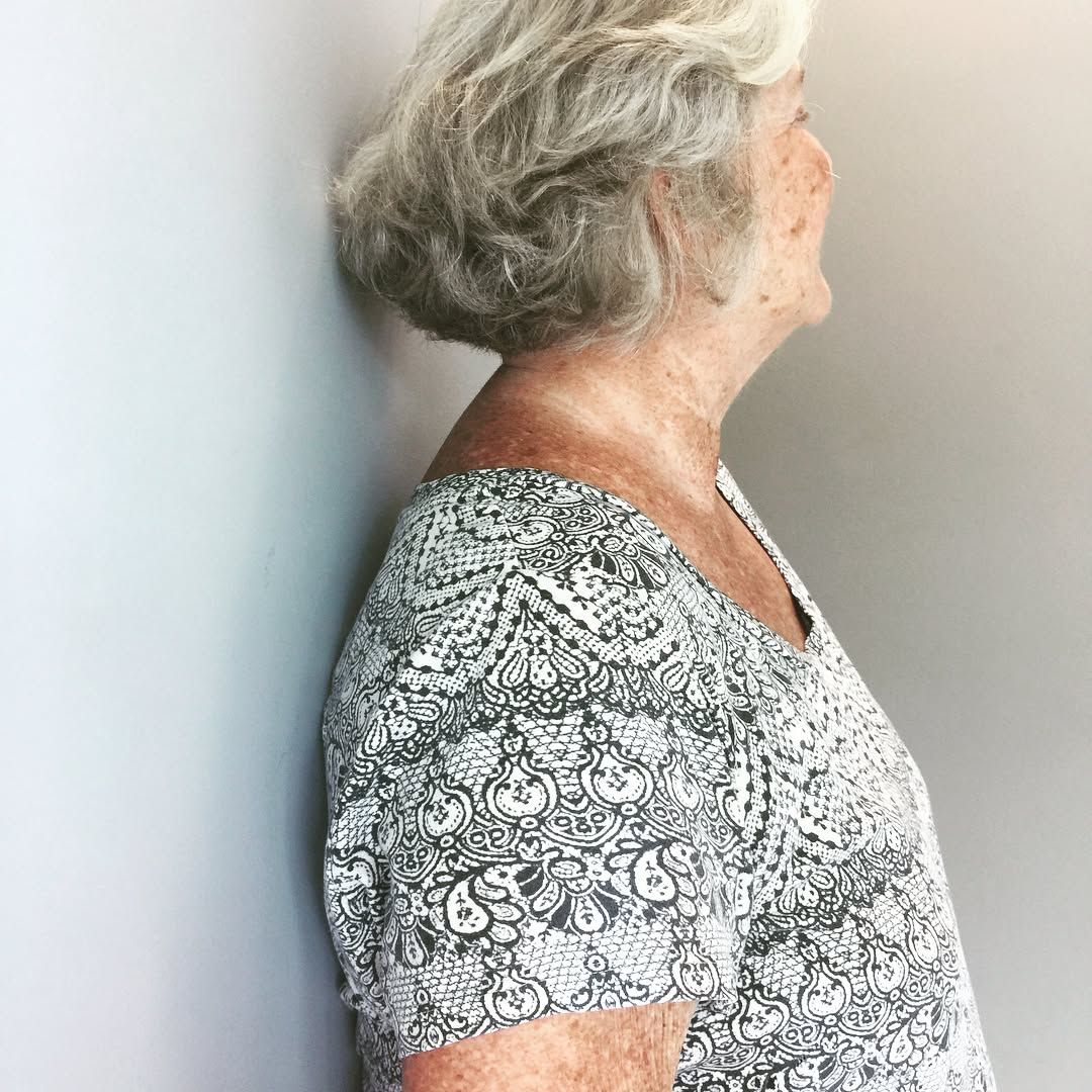 Senior woman with short grey hair leans against a plain wall, wearing a patterned blouse.