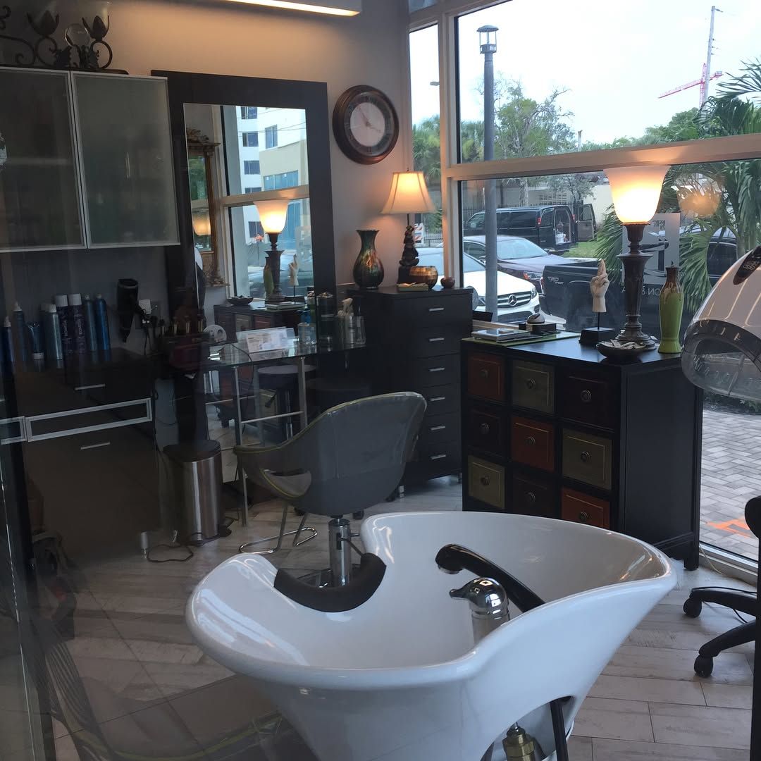 Salon interior with a white sink, mirror, and styling stations near a window.