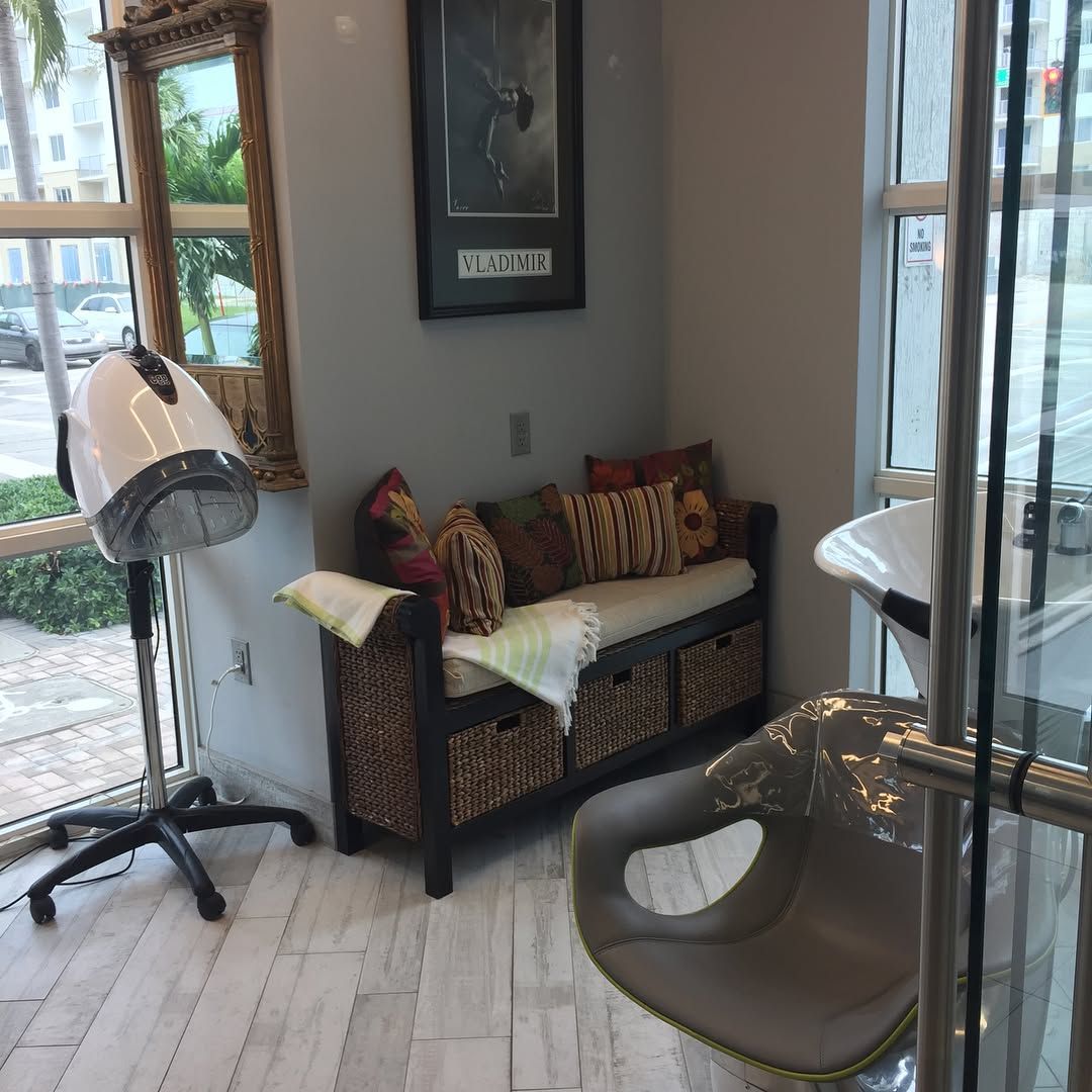 Hair salon corner: bench with pillows, dryer, chair, and a framed picture, near a window.