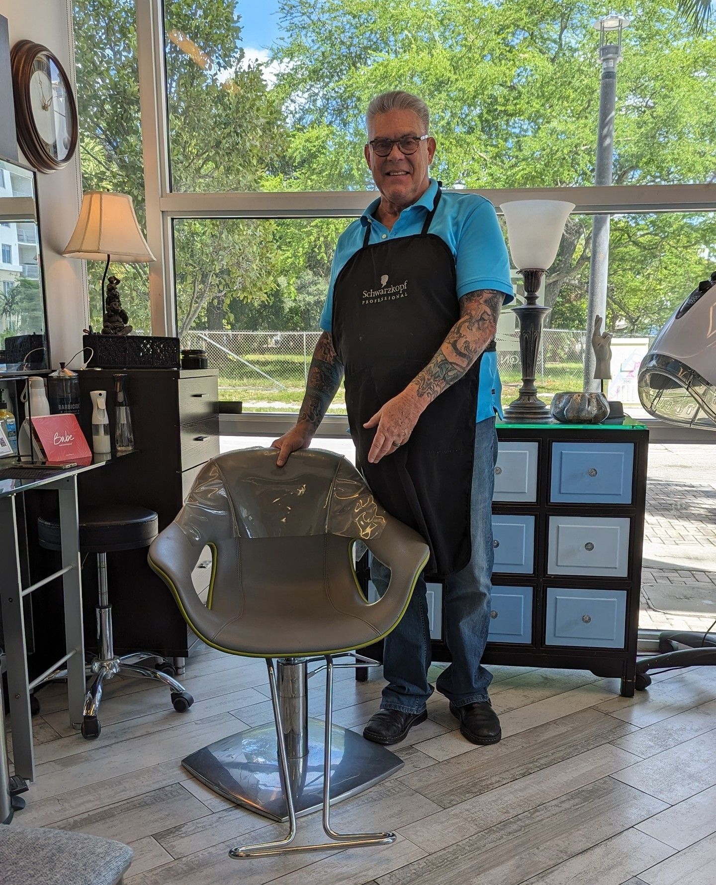 Man in a salon, wearing an apron, standing by a modern gray salon chair; window in the background.