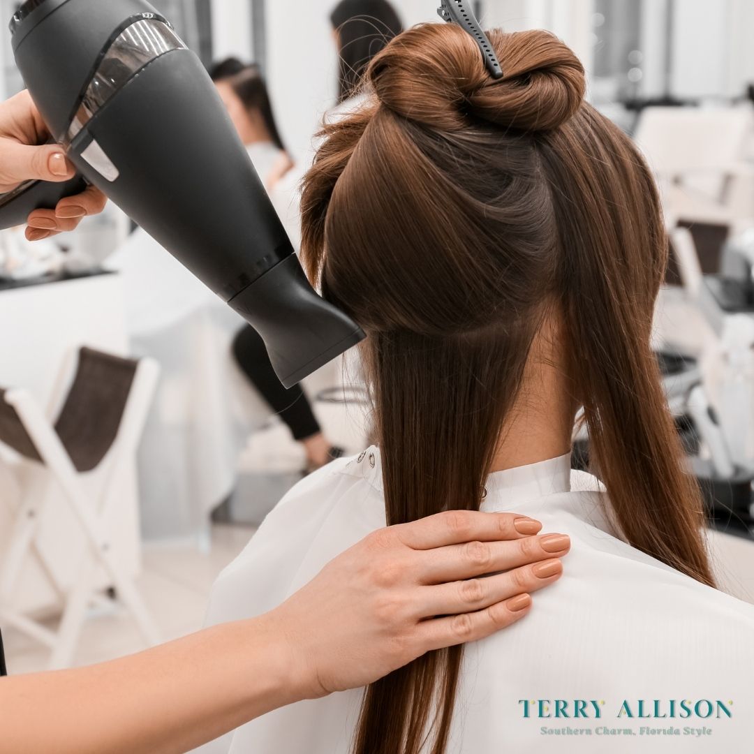 Hairdresser blow-drying a client's long brown hair in a salon setting.