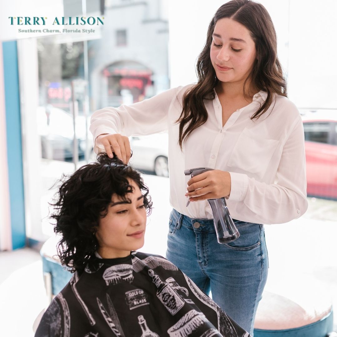 Hairdresser cutting a client's hair indoors. Client is seated, wearing a cape. Hairdresser is holding a spray bottle.