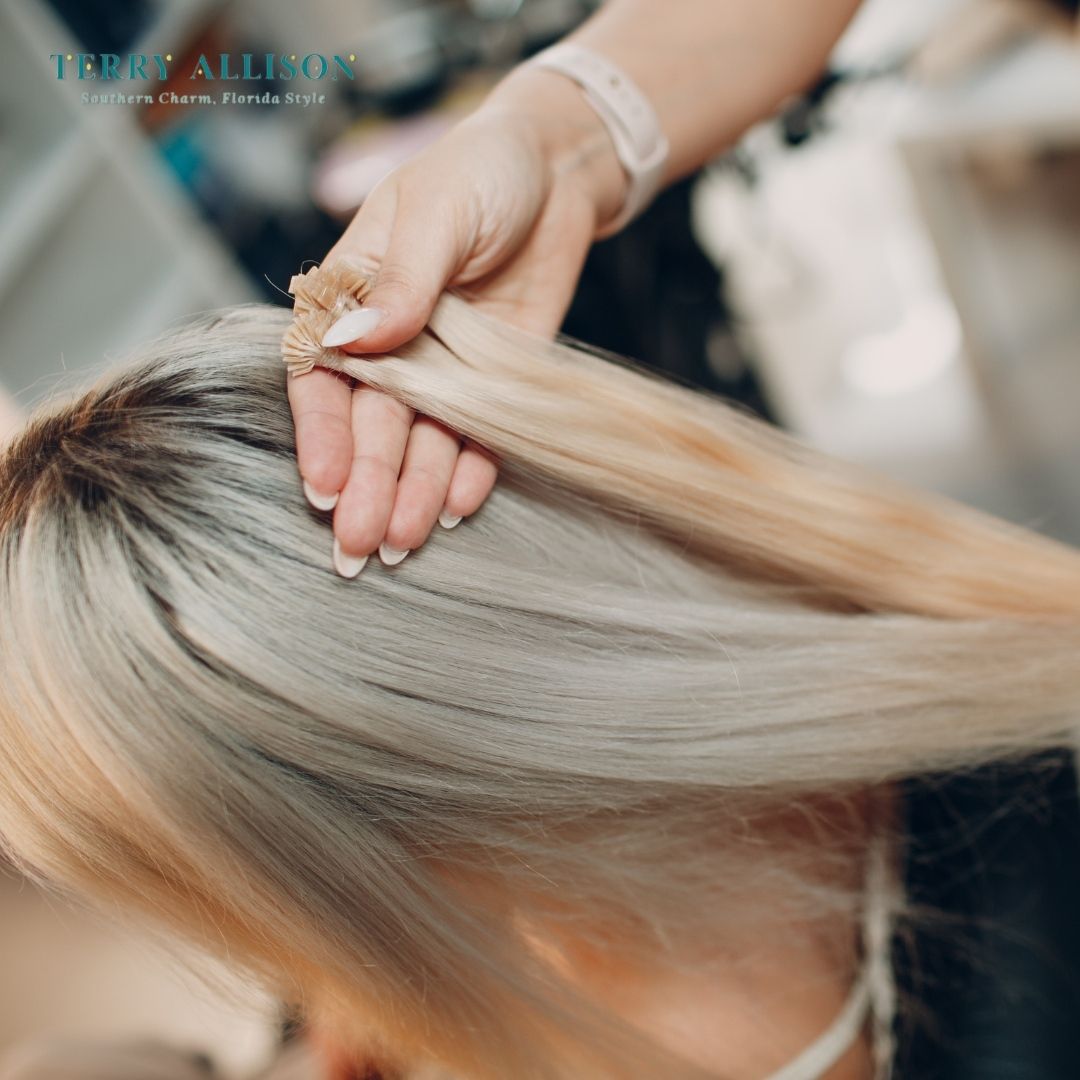 A stylist holding blonde hair extensions, adding to a client’s natural blonde hair at a salon.