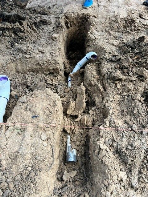 Trench dug in dirt revealing plumbing pipes; a section of white PVC pipe and metal fixture are visible.