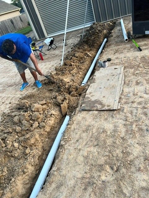 Man digging a trench for pipe installation near a building. Earth-toned setting.
