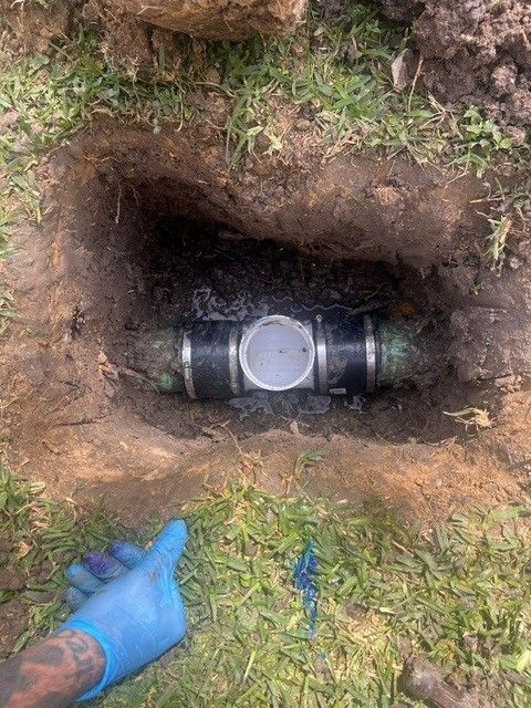 A hole in the ground reveals a black pipe with a white fitting. A gloved hand gives a thumbs up.