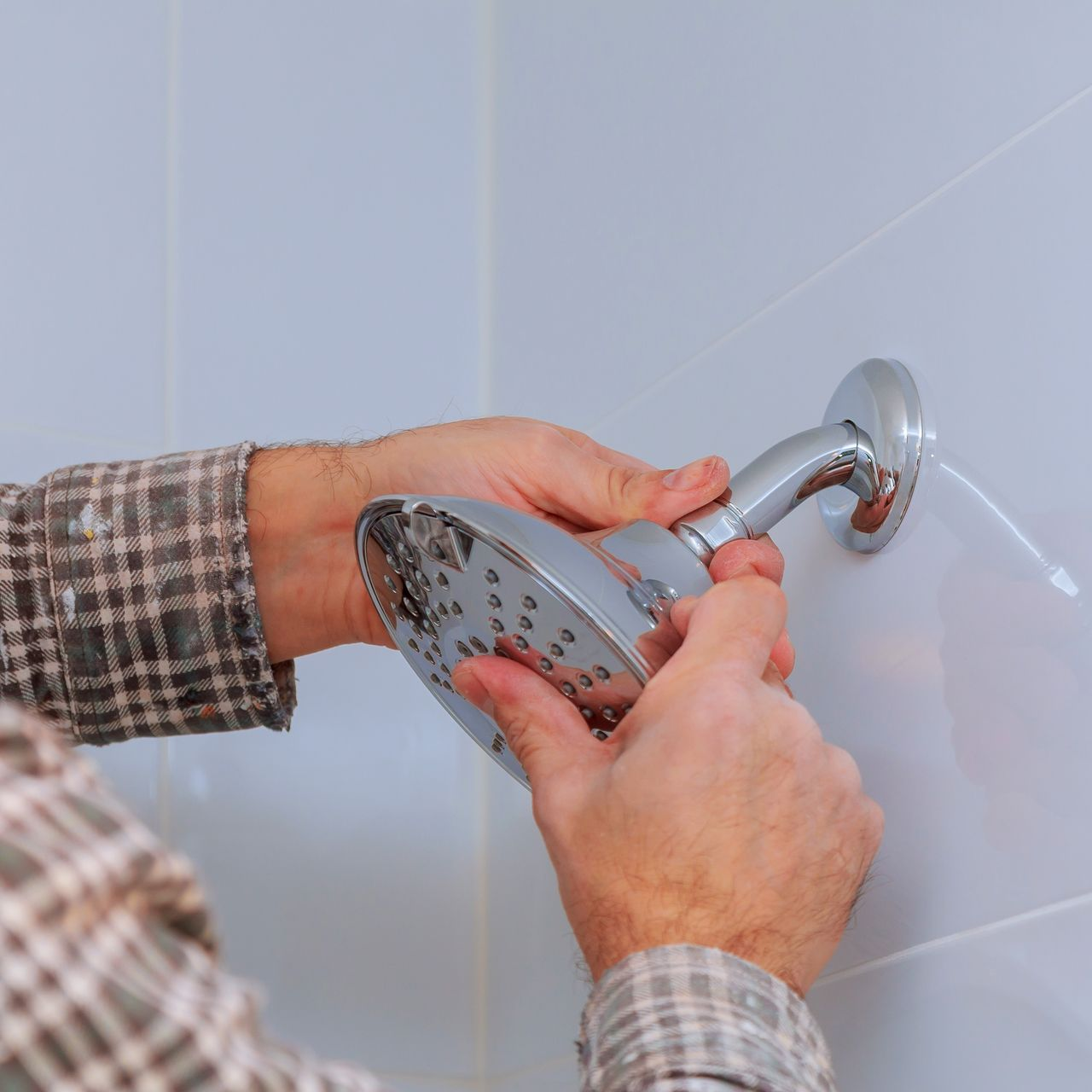 Person installing a chrome showerhead on a white-tiled wall, wearing a plaid shirt.