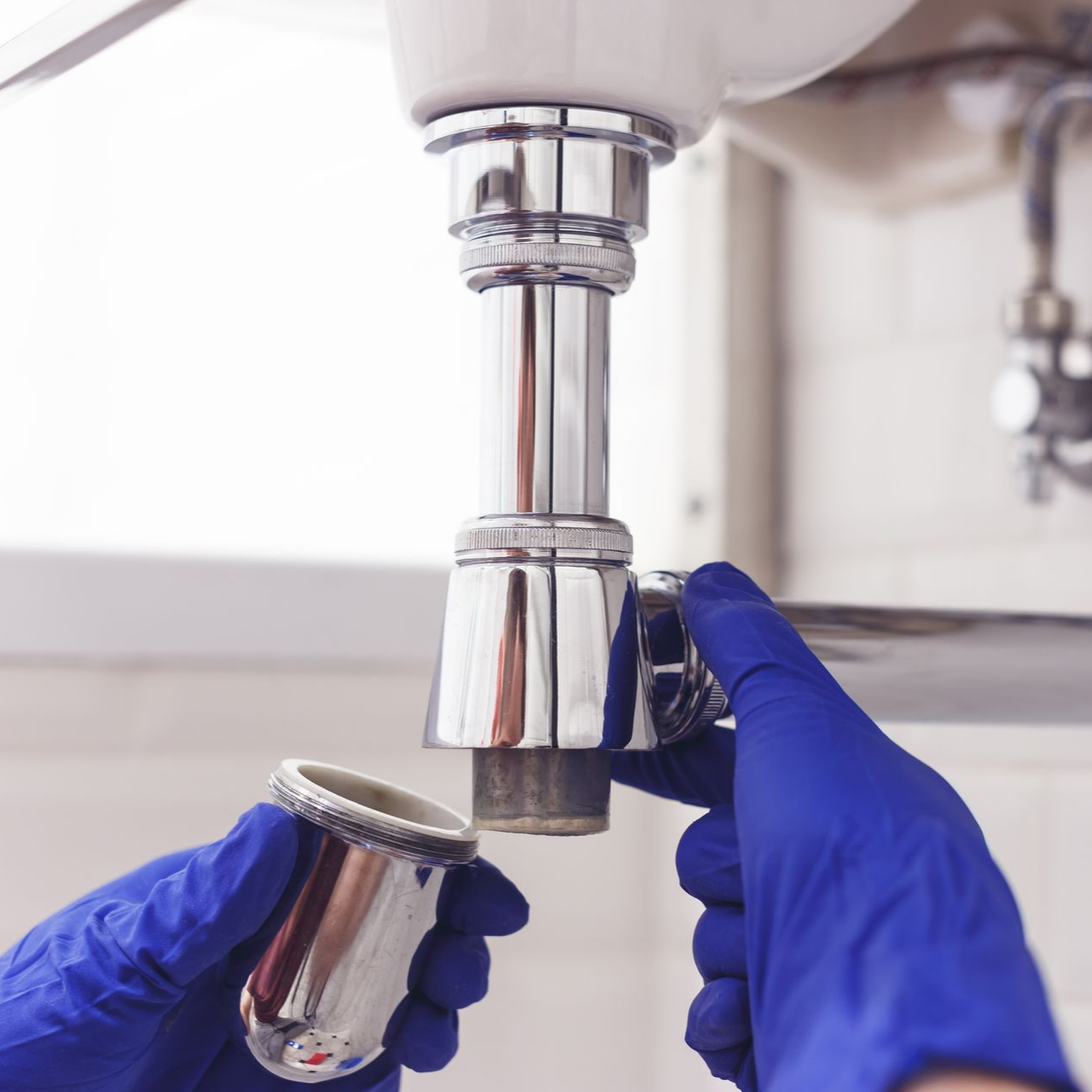 Hands in blue gloves unscrew a drain trap beneath a sink.