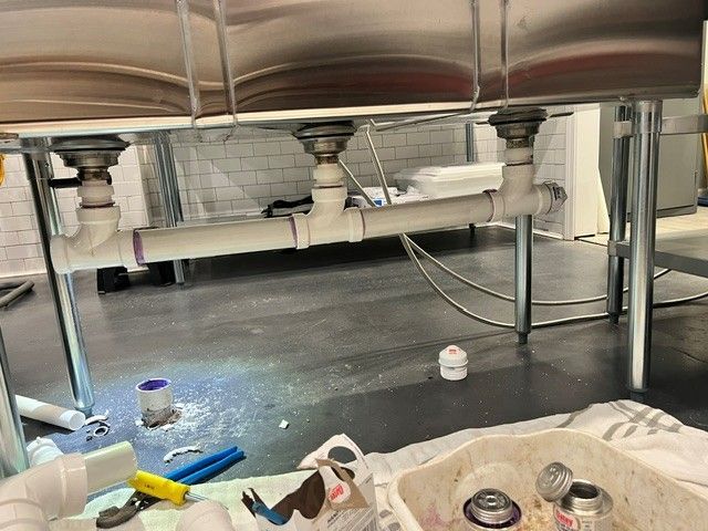 Under-sink plumbing with three drains connected by white PVC pipes. A toolbox and supplies lie on the floor.