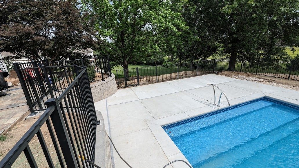 A pool with blue water, concrete deck, and black fence; trees in the background.