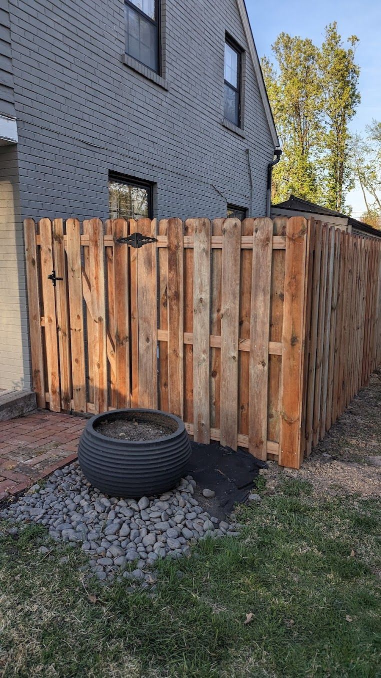 Wooden fence surrounding a backyard area with a large tire planter on the ground, next to the side of a brick house.