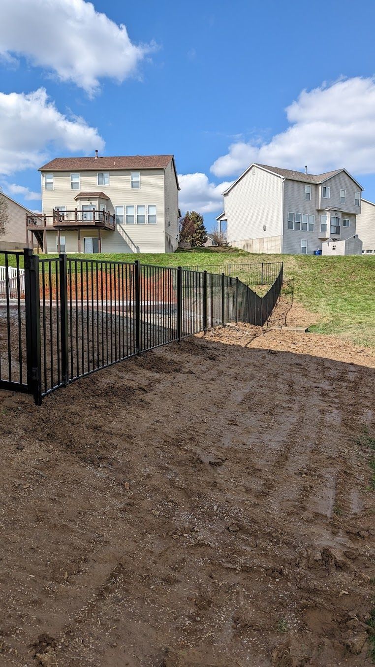 Black metal fence on sloped yard, with houses in the background, under a blue sky with clouds.