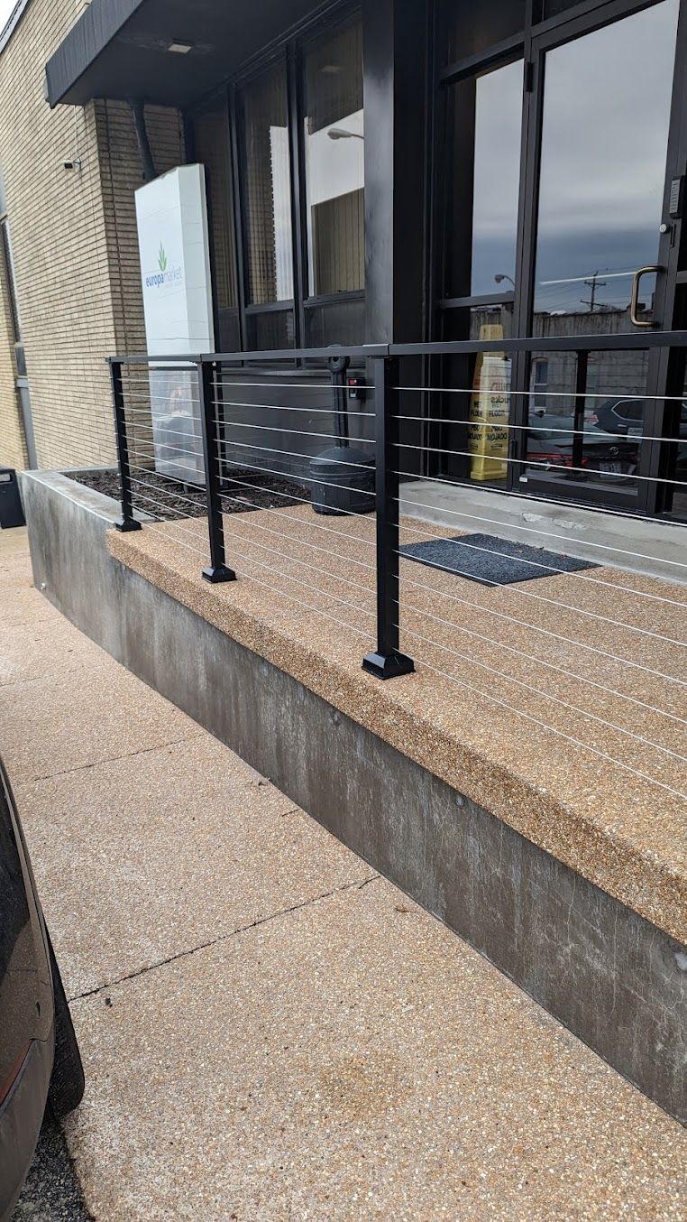 Black metal railing with horizontal cables on a brown concrete platform outside a building with large windows.