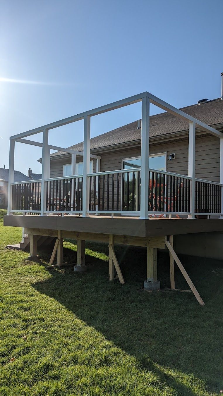 Deck with white pergola, black railing, supported by wooden posts on concrete blocks, set in a grassy yard.