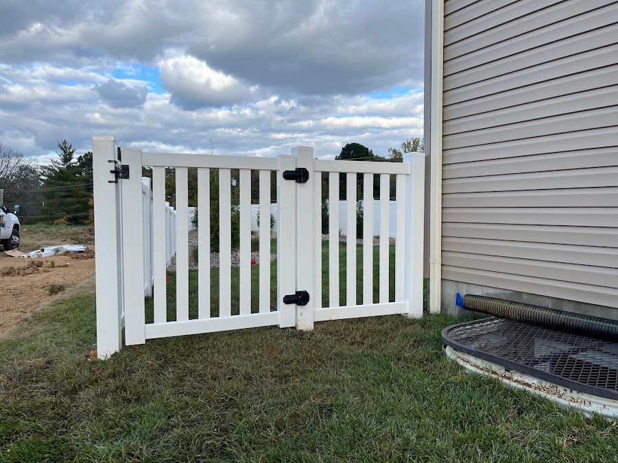 White picket fence gate next to a building's siding, set on a grassy lawn under a cloudy sky.