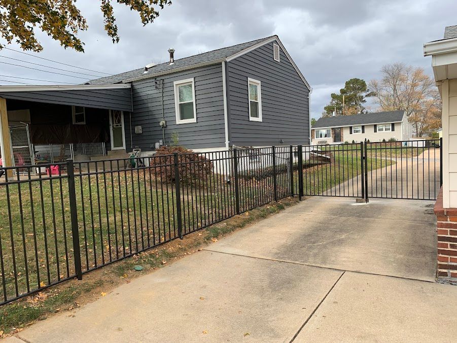 Black metal fence along a driveway, leading to a house with gray siding on a cloudy day.