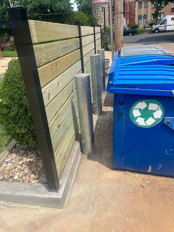 Blue recycling bin next to gray posts and a wooden fence on a sidewalk.