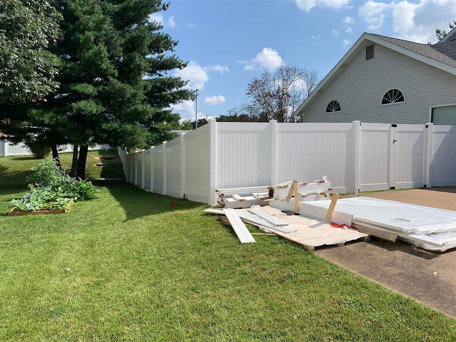 White vinyl fence with materials on the ground in a residential backyard on a sunny day.