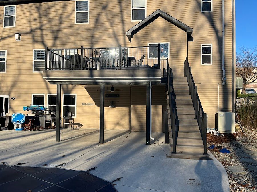 Deck with stairs, black railing, over a concrete patio. Beige siding on the building.