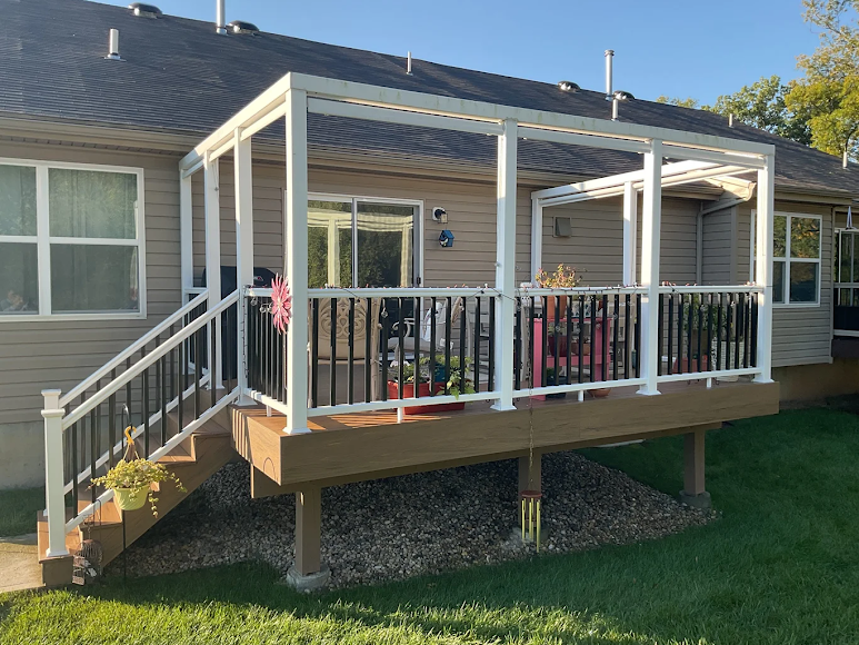 Backyard deck with white pergola, black railing, and steps, next to a tan house.