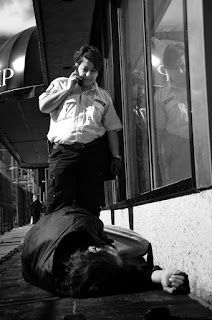 A black and white photo of a security guard talking on a cell phone, about to arrest a sleeping vagrant for the crime of being poor.