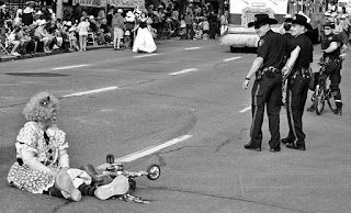 A black and white photo of a clown sitting on the side of the road after falling off their bicyle as police in cowboy hats look one.