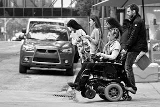 A man is helping to push a women in a wheel chair across a street crosswalk, while other pedestrians wait to cross behind them in the frame