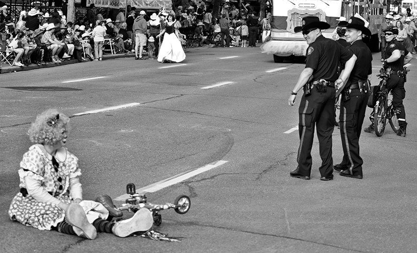 A black and white photo of a woman dressed as a clown sitting on the side of the road next to the tiny bicycle she fell off of. Police in Cowboy hats look on.