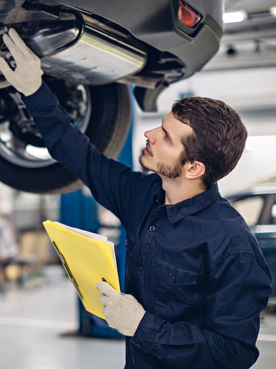 Mechanic inspects car underside, wearing gloves and holding a clipboard in a garage. | Railside Auto Service