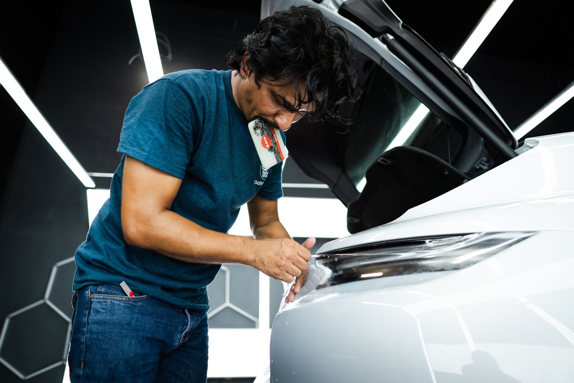 Man in blue shirt and jeans examining a white car's headlight.