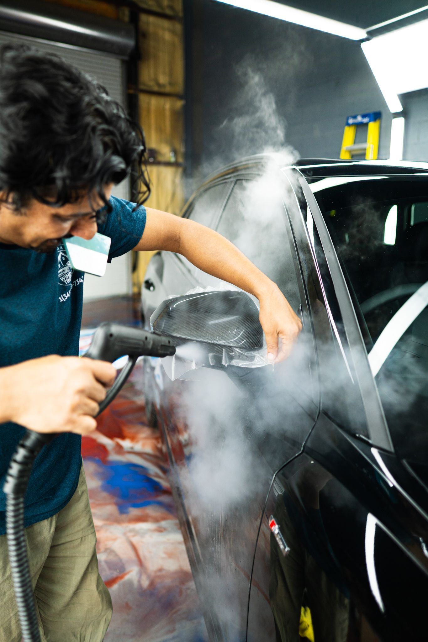 Man using a steam cleaner on a black car's side mirror, creating steam.