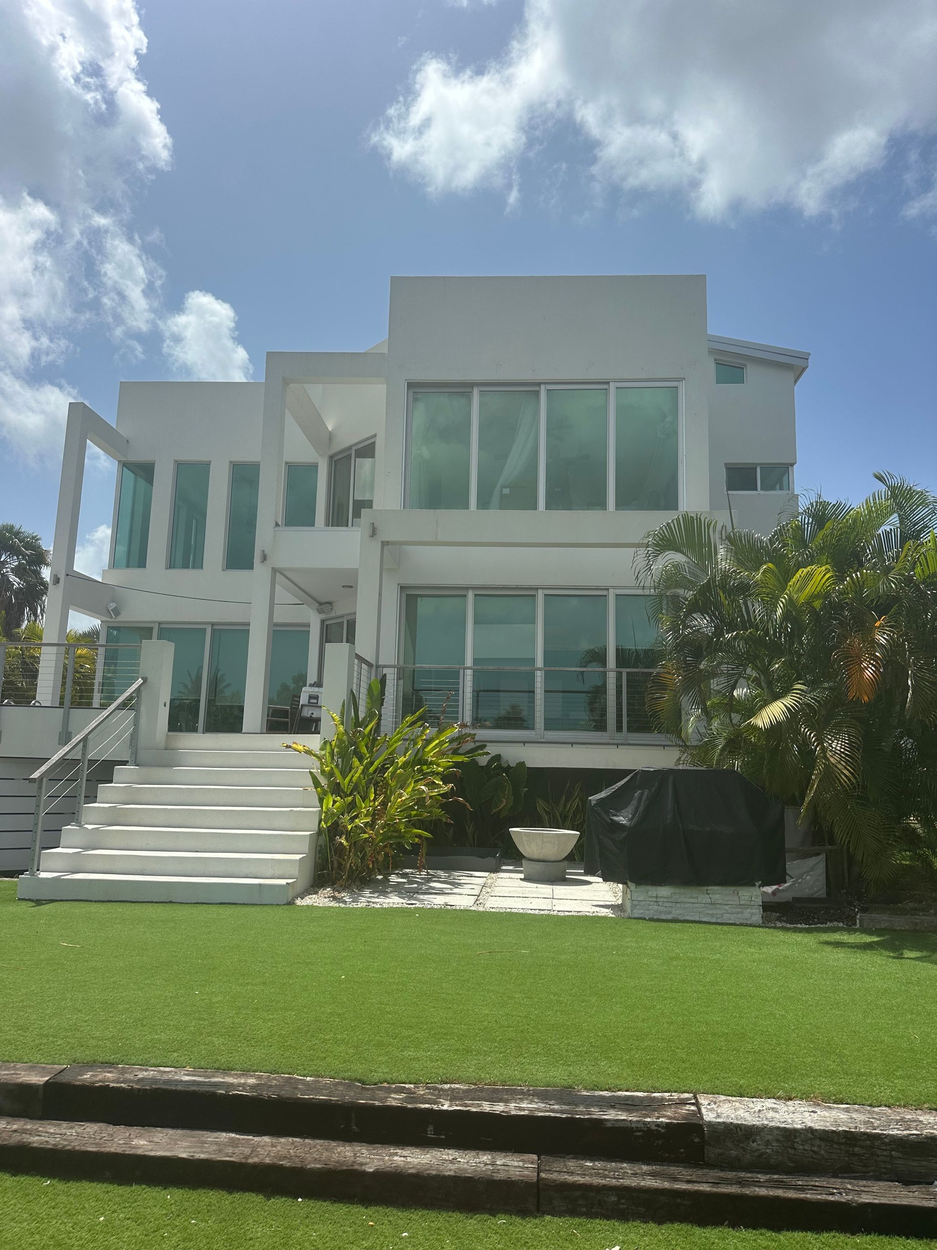 White modern waterfront house with large windows, stairs, and green grass.