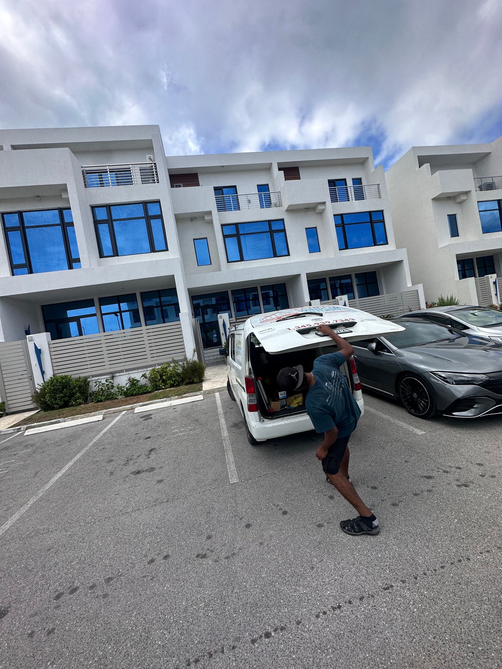 Person unloading groceries from a van in a parking lot of modern white buildings with blue-tinted windows.