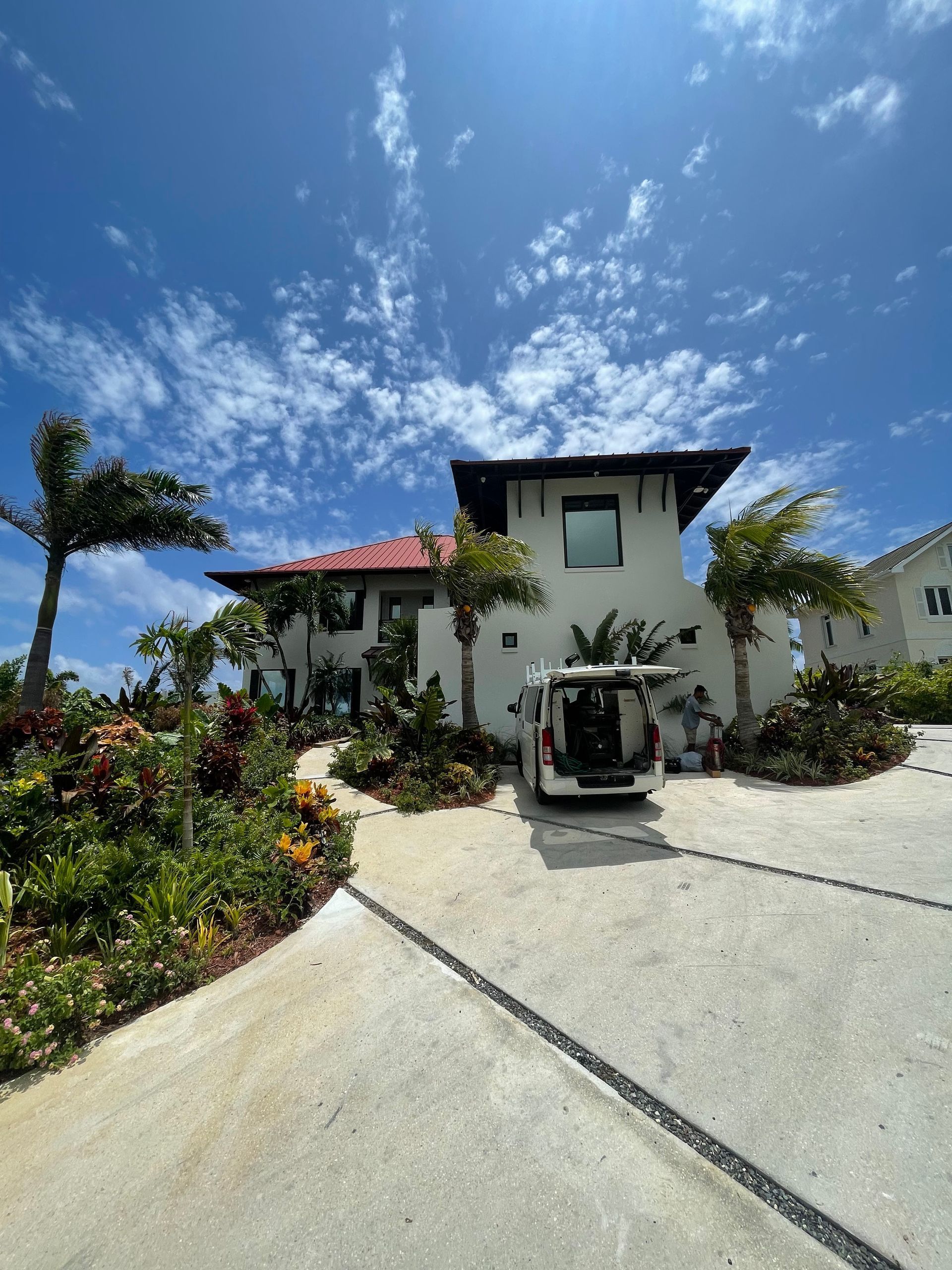 White house with red roof, palm trees, and a van in the driveway under a blue sky with clouds.